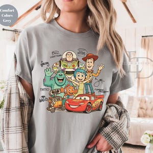 Puede incluir: Una camiseta gris de manga corta con un estampado gr&aacute;fico de personajes animados que incluye a Buzz Lightyear, Woody, Sulley, Nemo, Joy, Wall-E y Lightning McQueen, con sus nombres escritos en letra cursiva negra alrededor de las figuras.