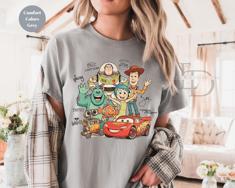Puede incluir: Una camiseta gris de manga corta con un estampado gr&aacute;fico de personajes animados que incluye a Buzz Lightyear, Woody, Sulley, Nemo, Joy, Wall-E y Lightning McQueen, con sus nombres escritos en letra cursiva negra alrededor de las figuras.