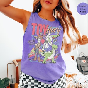 Puede incluir: Una camiseta sin mangas morada con la inscripci&oacute;n "Toy Story" y los personajes Woody, Buzz Lightyear y Rex. Tambi&eacute;n incluye el texto "Since 1995". La camiseta es de color violeta.