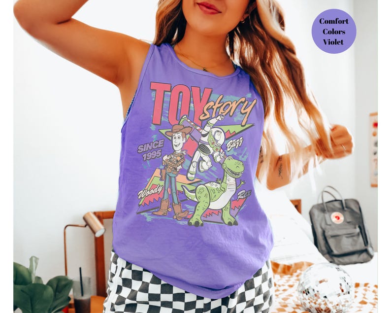 Puede incluir: Una camiseta sin mangas morada con la inscripci&oacute;n "Toy Story" y los personajes Woody, Buzz Lightyear y Rex. Tambi&eacute;n incluye el texto "Since 1995". La camiseta es de color violeta.