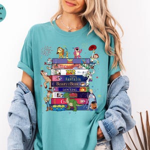 May include: A teal Comfort Colors t-shirt featuring a stack of VHS tapes with Disney movie titles and cartoon characters. The design includes characters from Toy Story, Beauty and the Beast, and The Lion King.