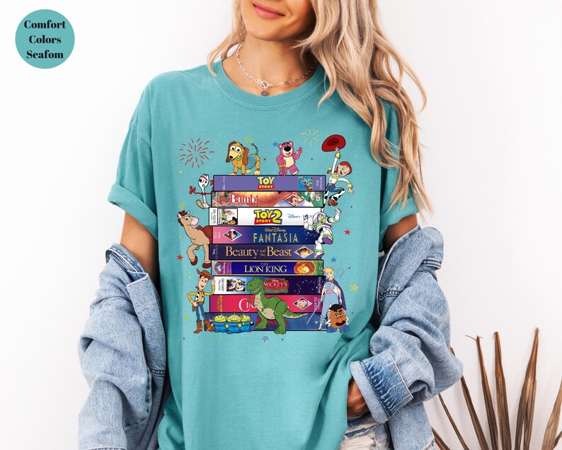 May include: A teal Comfort Colors t-shirt featuring a stack of VHS tapes with Disney movie titles and cartoon characters. The design includes characters from Toy Story, Beauty and the Beast, and The Lion King.