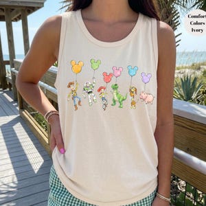 May include: Ivory tank top featuring a colorful graphic of cartoon characters holding Mickey Mouse-shaped balloons. The characters include a cowboy, a space ranger, a cowgirl, a green dinosaur, a dog, and a pig. The tank top is made of a soft material.