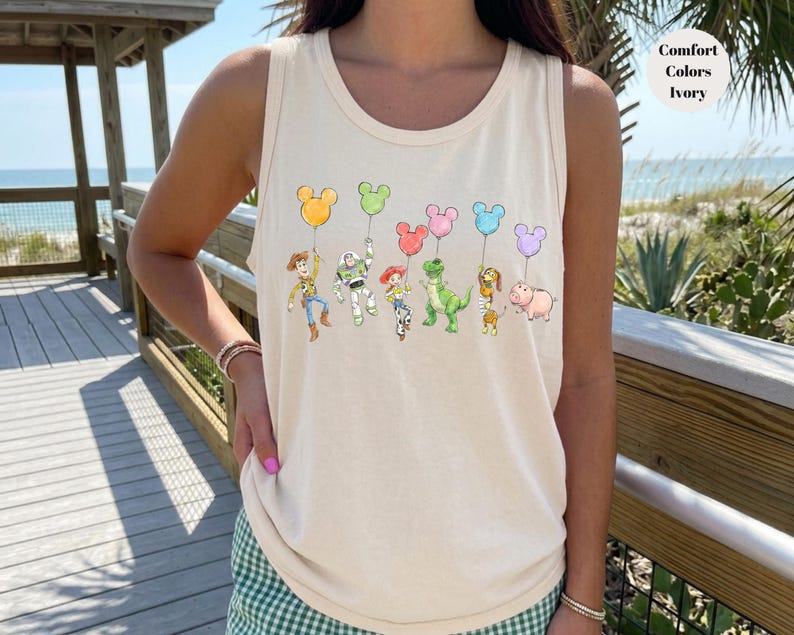 May include: Ivory tank top featuring a colorful graphic of cartoon characters holding Mickey Mouse-shaped balloons. The characters include a cowboy, a space ranger, a cowgirl, a green dinosaur, a dog, and a pig. The tank top is made of a soft material.