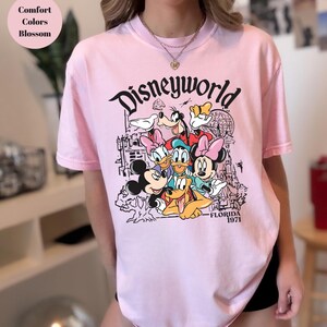 May include: A pale pink t-shirt with a graphic of Disney characters, including Mickey Mouse, Donald Duck, and Goofy. The word "Disneyworld" is printed above the characters, with "Florida 1971" below.