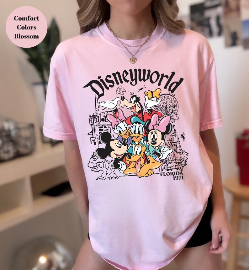 May include: A pale pink t-shirt with a graphic of Disney characters, including Mickey Mouse, Donald Duck, and Goofy. The word "Disneyworld" is printed above the characters, with "Florida 1971" below.