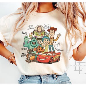 Puede incluir: Una camiseta de color crema con un estampado gr&aacute;fico de personajes de Disney y Pixar, incluidos Buzz Lightyear, Woody, Sulley, Joy, Nemo, Wall-E y Lightning McQueen, con sus nombres escritos en letra cursiva alrededor de las figuras.