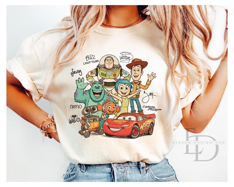 Puede incluir: Una camiseta de color crema con un estampado gr&aacute;fico de personajes de Disney y Pixar, incluidos Buzz Lightyear, Woody, Sulley, Joy, Nemo, Wall-E y Lightning McQueen, con sus nombres escritos en letra cursiva alrededor de las figuras.