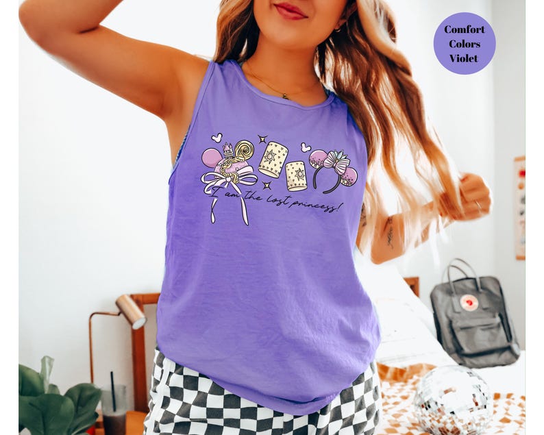 May include: A violet tank top with a graphic that includes a bow, candy, and mouse ears. The text "I am the lost princess!" is printed on the front. The words "Comfort Colors Violet" are in a circle in the upper right corner.