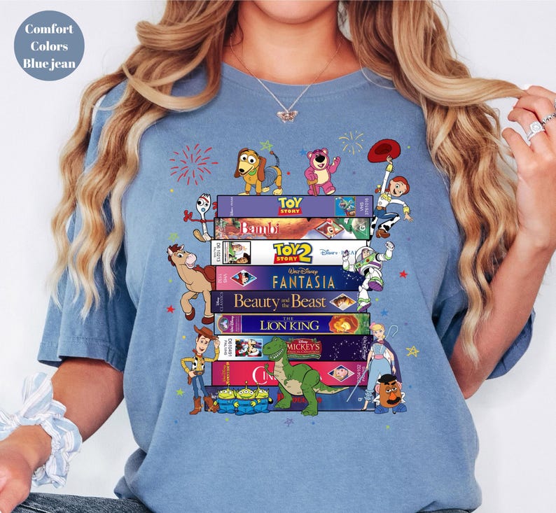 May include: Blue jean-colored t-shirt featuring a stack of VHS tapes with Disney movie titles and characters from Toy Story, Bambi, and Beauty and the Beast. The shirt has the words "Comfort Colors" and "Blue jean" printed on it.