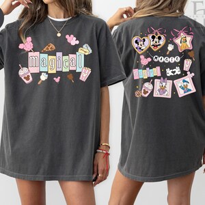 Op de afbeelding: Donkergrijs T-shirt met het woord "magical" in kleurrijke blokletters, omringd door cartoonvoedsel en karakterafbeeldingen. De achterkant van het shirt toont Disney-personages in hart- en stervormige frames, met het woord "magic" erboven.