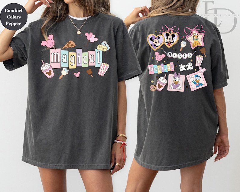 Op de afbeelding: Donkergrijs T-shirt met het woord "magical" in kleurrijke blokletters, omringd door cartoonvoedsel en karakterafbeeldingen. De achterkant van het shirt toont Disney-personages in hart- en stervormige frames, met het woord "magic" erboven.