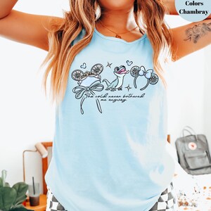 May include: Light blue tank top with a graphic featuring cartoon mouse ears, a bow, and a cartoon character. The text "The cold never bothered me anyway" is printed below the graphic. The tank top is made of chambray material.