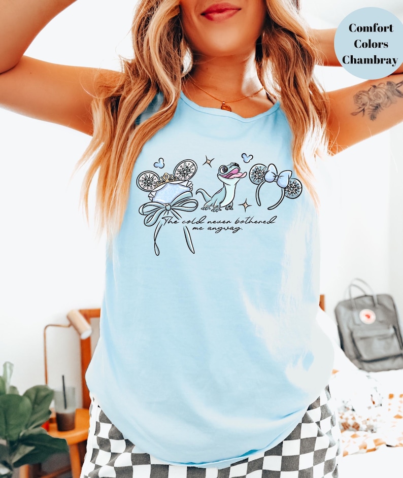 May include: Light blue tank top with a graphic featuring cartoon mouse ears, a bow, and a cartoon character. The text "The cold never bothered me anyway" is printed below the graphic. The tank top is made of chambray material.