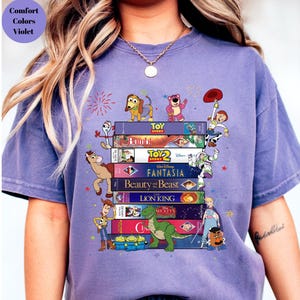 May include: A violet-colored t-shirt featuring a stack of Disney VHS tapes with characters from Toy Story, Bambi, Fantasia, Beauty and the Beast, The Lion King, and more. The shirt also has the text "Comfort Colors Violet".