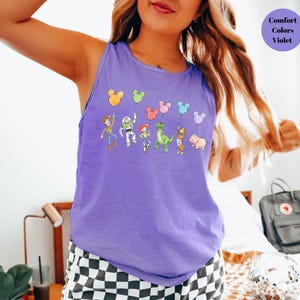 May include: A violet tank top featuring a cartoon design of characters holding balloons. The balloons are in the shape of Mickey Mouse heads. The text "Comfort Colors Violet" is in a circle on the upper right.