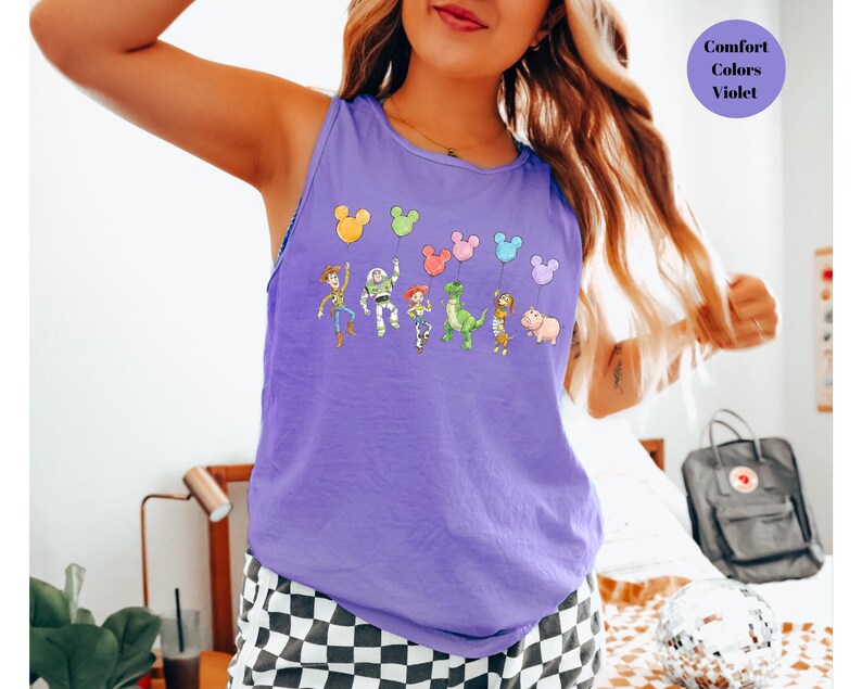 May include: A violet tank top featuring a cartoon design of characters holding balloons. The balloons are in the shape of Mickey Mouse heads. The text "Comfort Colors Violet" is in a circle on the upper right.