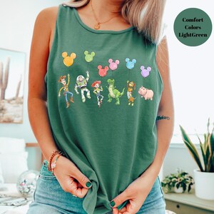 May include: A light green tank top featuring a colorful graphic of cartoon characters holding Mickey Mouse-shaped balloons. The characters include a cowboy, a space ranger, a cowgirl, a dinosaur, a monkey, and a pig. The text "Comfort Colors Light Green" is in the upper right corner.