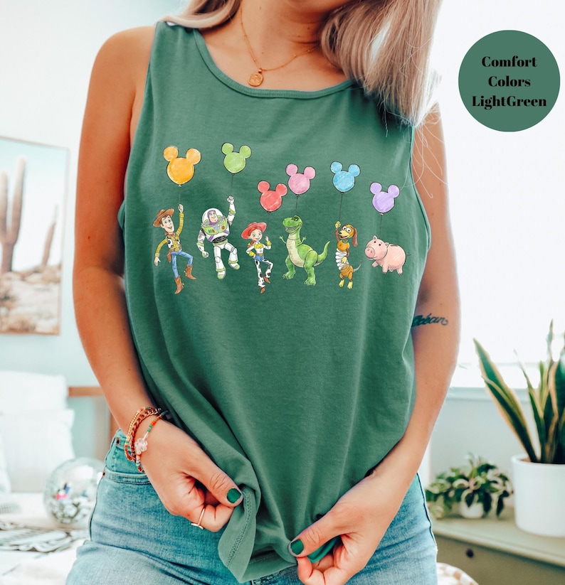 May include: A light green tank top featuring a colorful graphic of cartoon characters holding Mickey Mouse-shaped balloons. The characters include a cowboy, a space ranger, a cowgirl, a dinosaur, a monkey, and a pig. The text "Comfort Colors Light Green" is in the upper right corner.