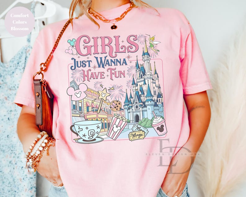 May include: Pink t-shirt with the phrase "GIRLS JUST WANNA HAVE FUN" and a colorful graphic featuring a castle, Mickey Mouse, popcorn, and a drink. The shirt has short sleeves and a crew neck.
