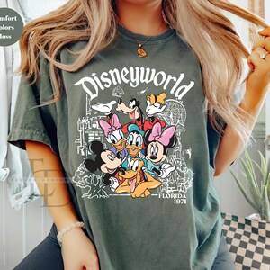 May include: Moss green t-shirt with a Disneyworld graphic featuring Mickey Mouse, Minnie Mouse, Donald Duck, Daisy Duck, Goofy, and Pluto. The text "Disneyworld Florida 1971" is printed in white. The shirt has a crew neck.