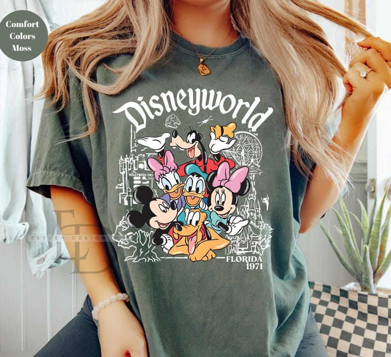 May include: Moss green t-shirt with a Disneyworld graphic featuring Mickey Mouse, Minnie Mouse, Donald Duck, Daisy Duck, Goofy, and Pluto. The text "Disneyworld Florida 1971" is printed in white. The shirt has a crew neck.