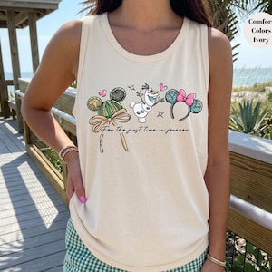 May include: Ivory tank top with a whimsical design featuring Mickey Mouse ears, Olaf, and Minnie Mouse ears. The text "For the first time in forever" is written below the characters. The tank top is a Comfort Colors brand.