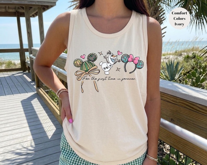 May include: Ivory tank top with a whimsical design featuring Mickey Mouse ears, Olaf, and Minnie Mouse ears. The text "For the first time in forever" is written below the characters. The tank top is a Comfort Colors brand.