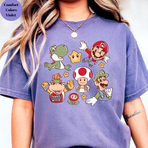 May include: A violet Comfort Colors t-shirt featuring a colorful graphic of various characters from the Super Mario video game series. The design includes Mario, Luigi, Yoshi, Bowser, Toad, and other elements.