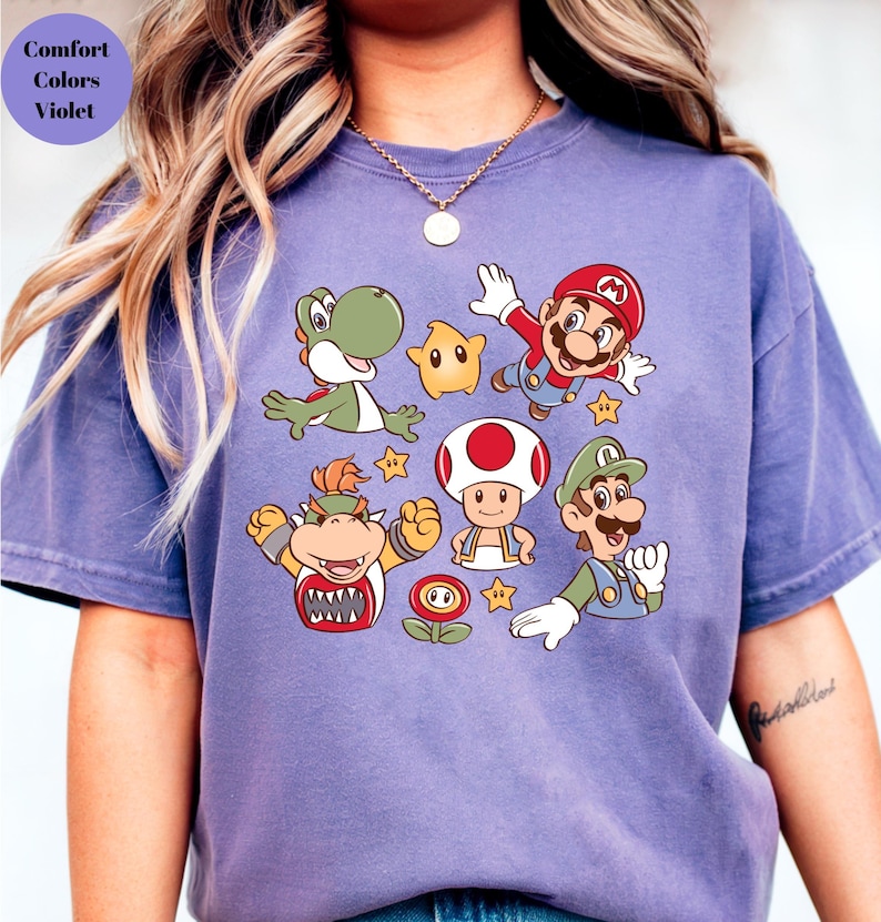 May include: A violet Comfort Colors t-shirt featuring a colorful graphic of various characters from the Super Mario video game series. The design includes Mario, Luigi, Yoshi, Bowser, Toad, and other elements.