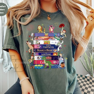 May include: A moss green t-shirt featuring a stack of VHS tapes with classic Disney movie titles and characters. The design includes characters from Toy Story, Bambi, Beauty and the Beast, and The Lion King. The shirt has the words "Comfort Colors Moss" in the upper left corner.