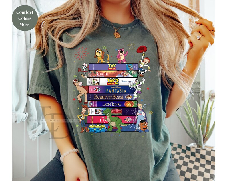 May include: A moss green t-shirt featuring a stack of VHS tapes with classic Disney movie titles and characters. The design includes characters from Toy Story, Bambi, Beauty and the Beast, and The Lion King. The shirt has the words "Comfort Colors Moss" in the upper left corner.