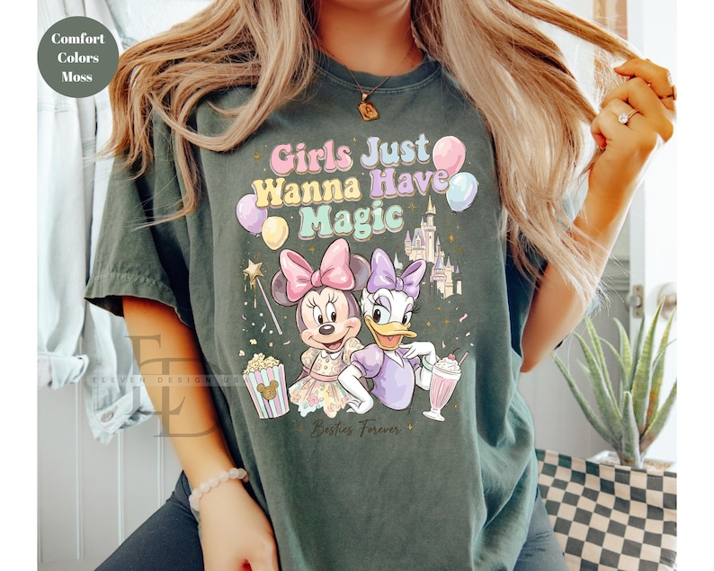 Puede incluir: Camiseta verde oscuro con el texto "Girls Just Wanna Have Magic" e ilustraciones de Minnie Mouse, Daisy Duck, un castillo y globos. La camiseta tambi&eacute;n presenta palomitas de ma&iacute;z y un batido.