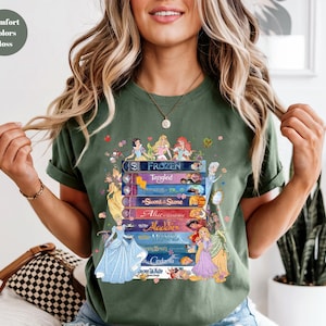 Puede incluir: Camiseta verde musgo con una pila de libros de cuentos de Disney con ilustraciones de princesas. Los t&iacute;tulos incluyen "Frozen", "Tangled", "Sword in the Stone", "Alice", "Aladdin", "Mermaid", "Beauty", "Cinderella" y "Snow White".