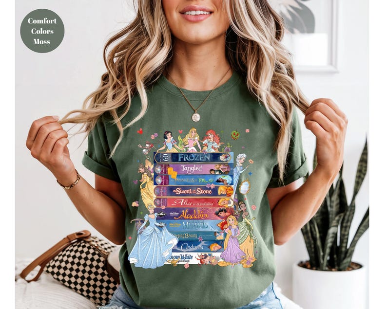 Puede incluir: Camiseta verde musgo con una pila de libros de cuentos de Disney con ilustraciones de princesas. Los t&iacute;tulos incluyen "Frozen", "Tangled", "Sword in the Stone", "Alice", "Aladdin", "Mermaid", "Beauty", "Cinderella" y "Snow White".