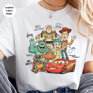 Puede incluir: Una camiseta blanca con un estampado gr&aacute;fico de varios personajes animados, incluidos Buzz Lightyear, Woody, Sulley, Joy, Nemo, Wall-E y Lightning McQueen, con sus nombres escritos en una fuente manuscrita estilizada junto a cada personaje.