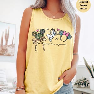 May include: Yellow tank top with a Disney-themed graphic. The design includes Mickey Mouse ears, Olaf the snowman, and the phrase "For the first time in forever." The top is a comfort color in butter yellow.
