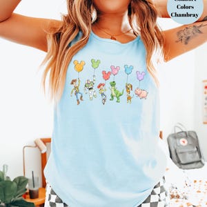 May include: Light blue tank top featuring a colorful graphic of cartoon characters holding Mickey Mouse-shaped balloons. The text "Comfort Colors Chambray" is in a light blue circle. The tank top is made of a soft, comfortable material.