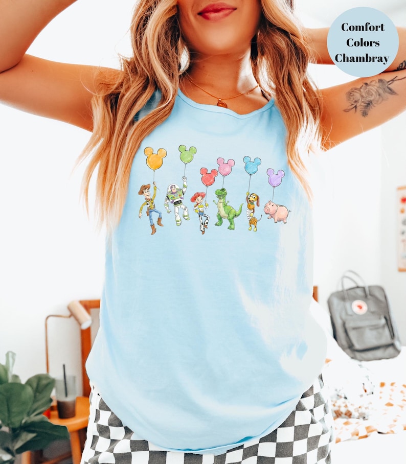May include: Light blue tank top featuring a colorful graphic of cartoon characters holding Mickey Mouse-shaped balloons. The text "Comfort Colors Chambray" is in a light blue circle. The tank top is made of a soft, comfortable material.