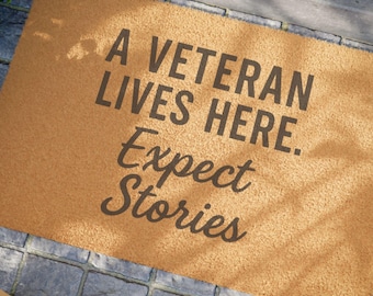 May include: A rectangular doormat with the text "A VETERAN LIVES HERE. Expect Stories" in dark gray lettering. The mat is a light brown color and sits on a gray stone surface.