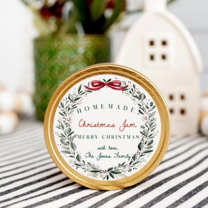 Pode incluir: Tampa redonda de frasco com borda dourada e rótulo que diz "Homemade Christmas Jam". O rótulo apresenta um desenho de coroa com um laço vermelho e o texto "Merry Christmas" e "with love, The Jones Family."