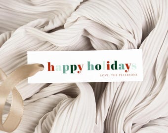 May include: A white gift tag with the words "happy holidays" in red, green, and light blue lettering. The tag is attached to a gold ribbon and rests on a textured, off-white fabric. The text "Love, The Petersons" is also on the tag.