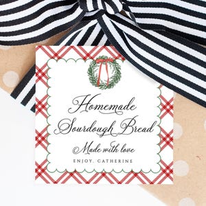 Może przedstawiać: Kwadratowa etykieta prezentowa z czerwoną i zieloną kratką oraz ząbkowanym brzegiem. Etykieta zawiera tekst "Homemade Sourdough Bread Made with love" i akwarelowy wieniec. Czarno-biała wstążka w paski jest zawiązana wokół prezentu.