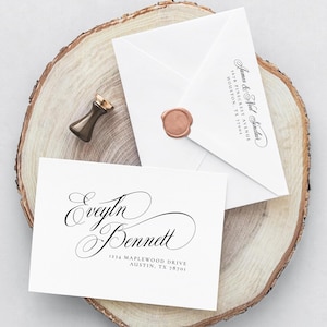 May include: A white invitation suite featuring elegant calligraphy. The suite includes a card with the names "Evelyn Bennett" and address, and an envelope sealed with a wax stamp. A gold-toned wax stamp tool rests on a wooden slice.
