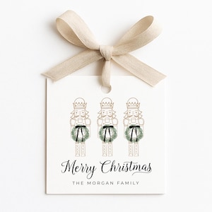 May include: A white gift tag with three illustrated nutcrackers, each wearing a green wreath and black bow. The tag is tied with a beige ribbon and features the text "Merry Christmas" and "The Morgan Family".