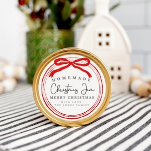 May include: A gold metal jar with a white label that says "Homemade Christmas Jam Merry Christmas With Love The Jones Family". The label has a red bow and a red circle around the text.