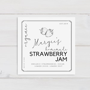 Op de afbeelding: Zwart-wit label met de tekst "Marquis' homemade STRAWBERRY JAM" en de tekst "ORGANIC STRAWBERRIES, SUGAR. LEMON JUICE. LEMON ZEST" eronder. Het label bevat ook een tekening van twee aardbeien en de tekst "EST. 2019" in de rechterbovenhoek.