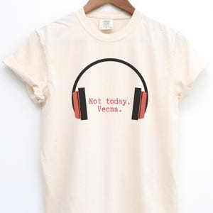 May include: Cream-colored t-shirt with a black and red headphone graphic. The text "Not today, Vecna." is printed in red. The shirt is hanging on a wooden hanger.