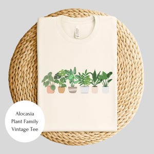 May include: A cream-colored t-shirt with a graphic of potted plants. The text "Alocasia Plant Family Vintage Tee" is printed on the bottom left corner of the shirt.
