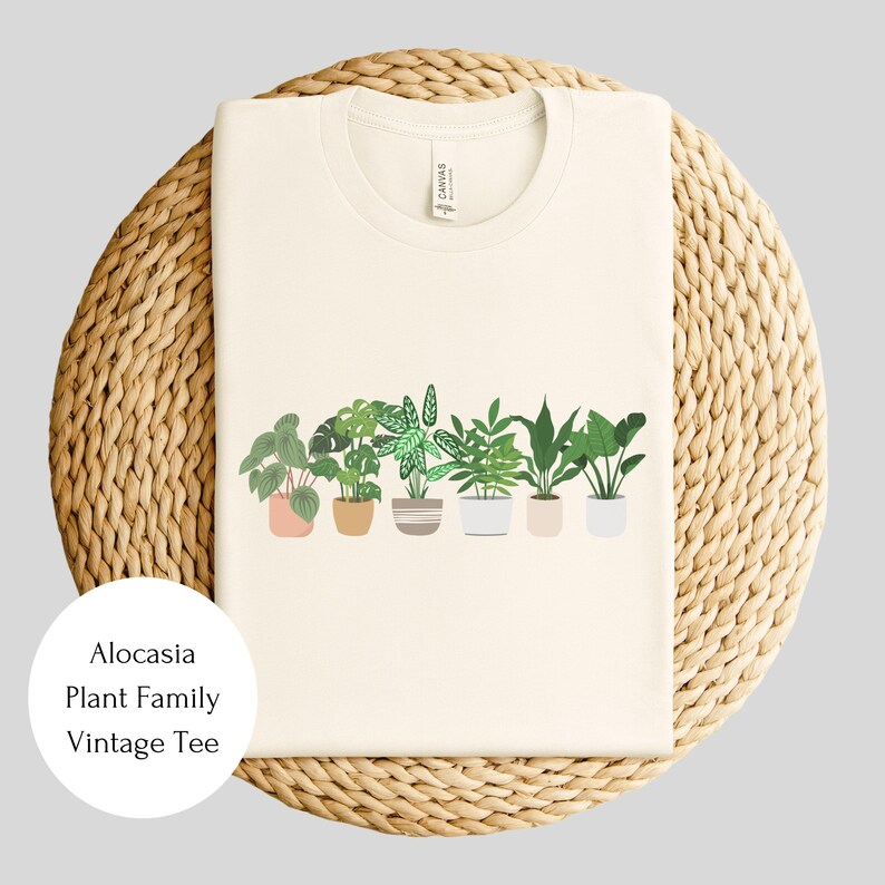 May include: A cream-colored t-shirt with a graphic of potted plants. The text "Alocasia Plant Family Vintage Tee" is printed on the bottom left corner of the shirt.